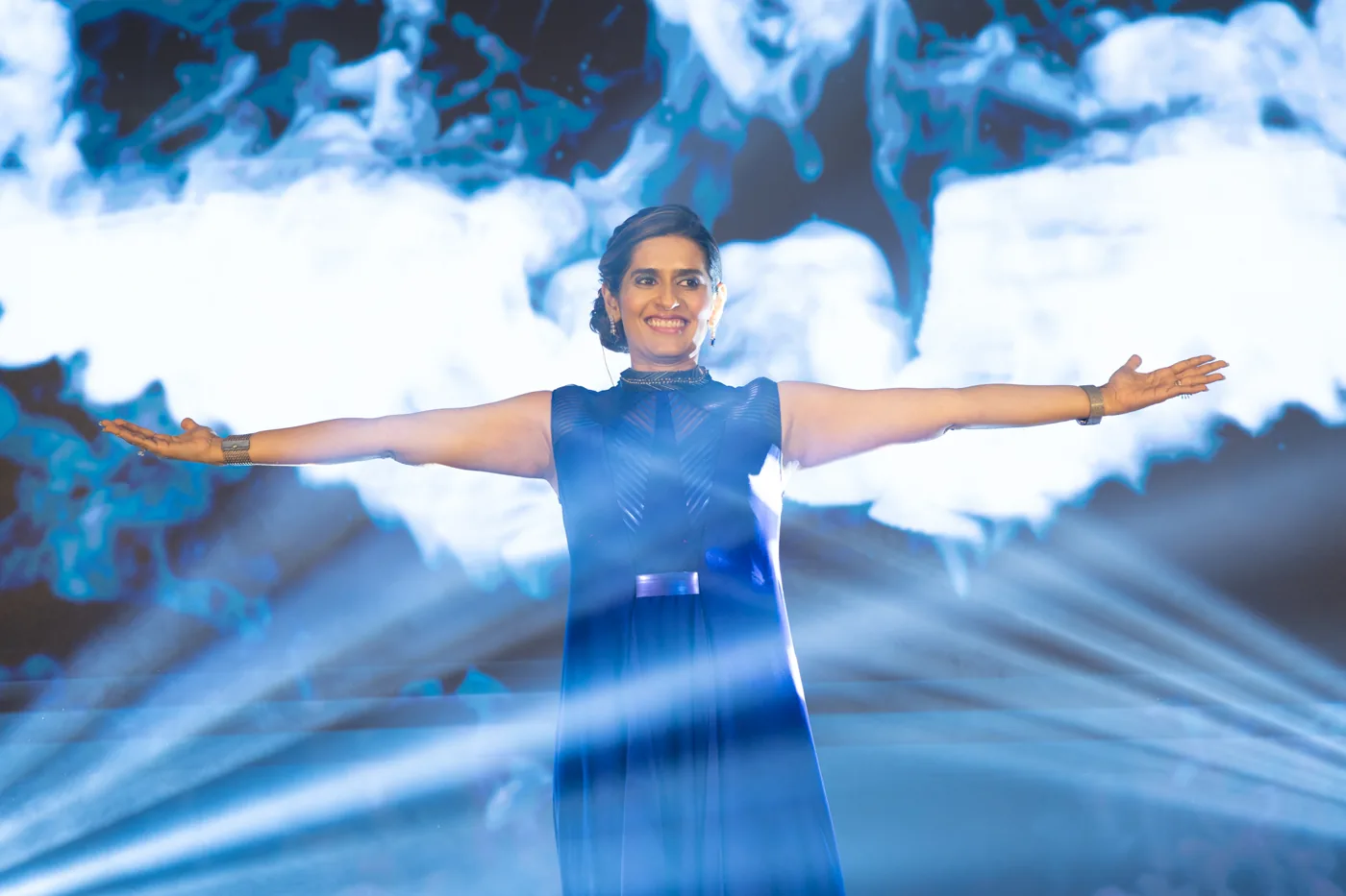 Ritu Agarwal on stage, arms open, addressing the community